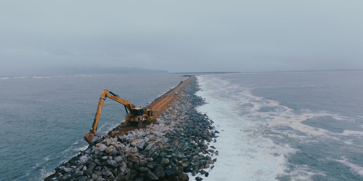An excavator places armor stone on the jetty as waves roll past.