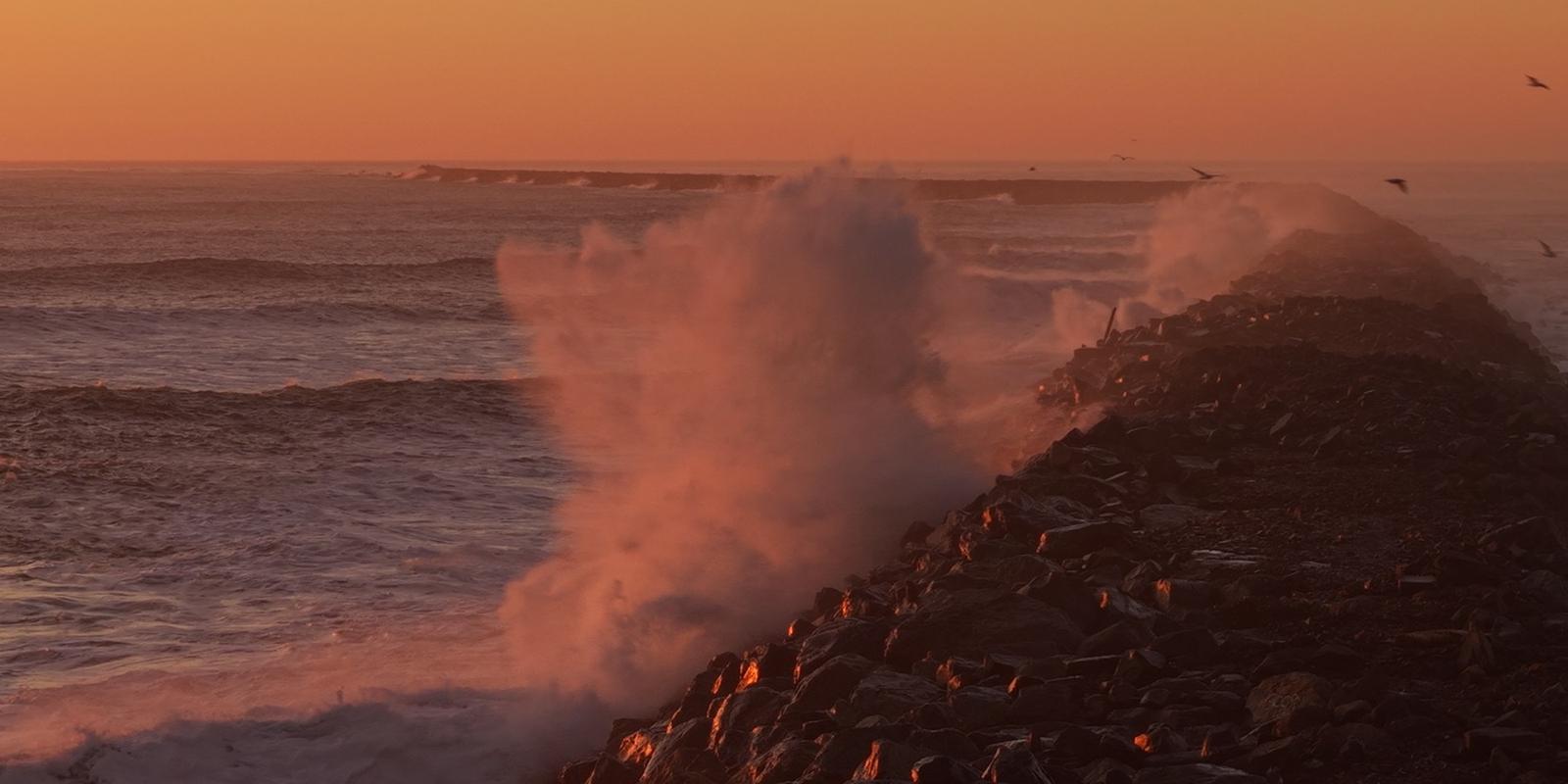 Surf explodes against the rock jetty at sunset.